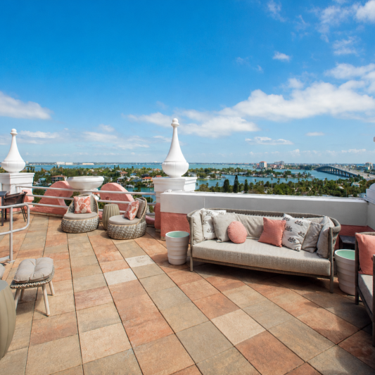 Rooftop terrace with beige wicker seating and coral cushions, overlooking the ocean and a coastal city under a blue sky.