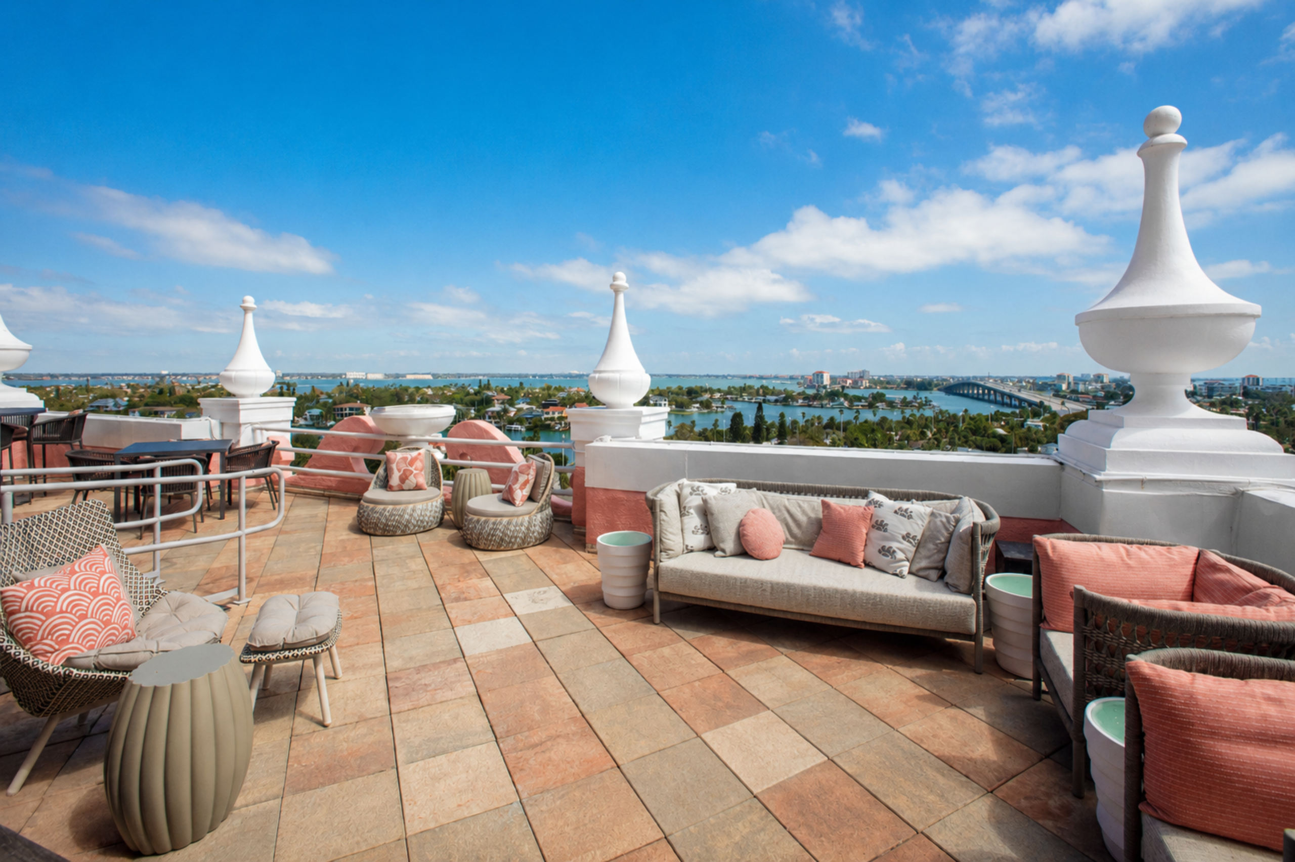 Rooftop terrace with beige wicker seating and coral cushions, overlooking the ocean and a coastal city under a blue sky.