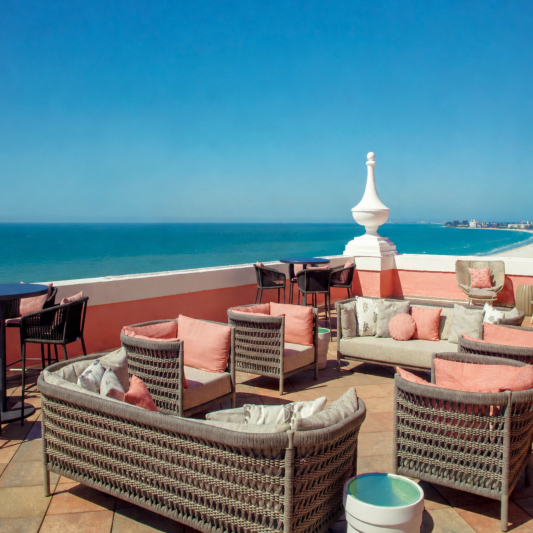 Seaside rooftop lounge with wicker chairs and pink cushions overlooking a blue ocean.