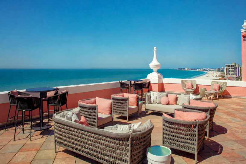 Seaside rooftop lounge with wicker chairs and pink cushions overlooking a blue ocean.
