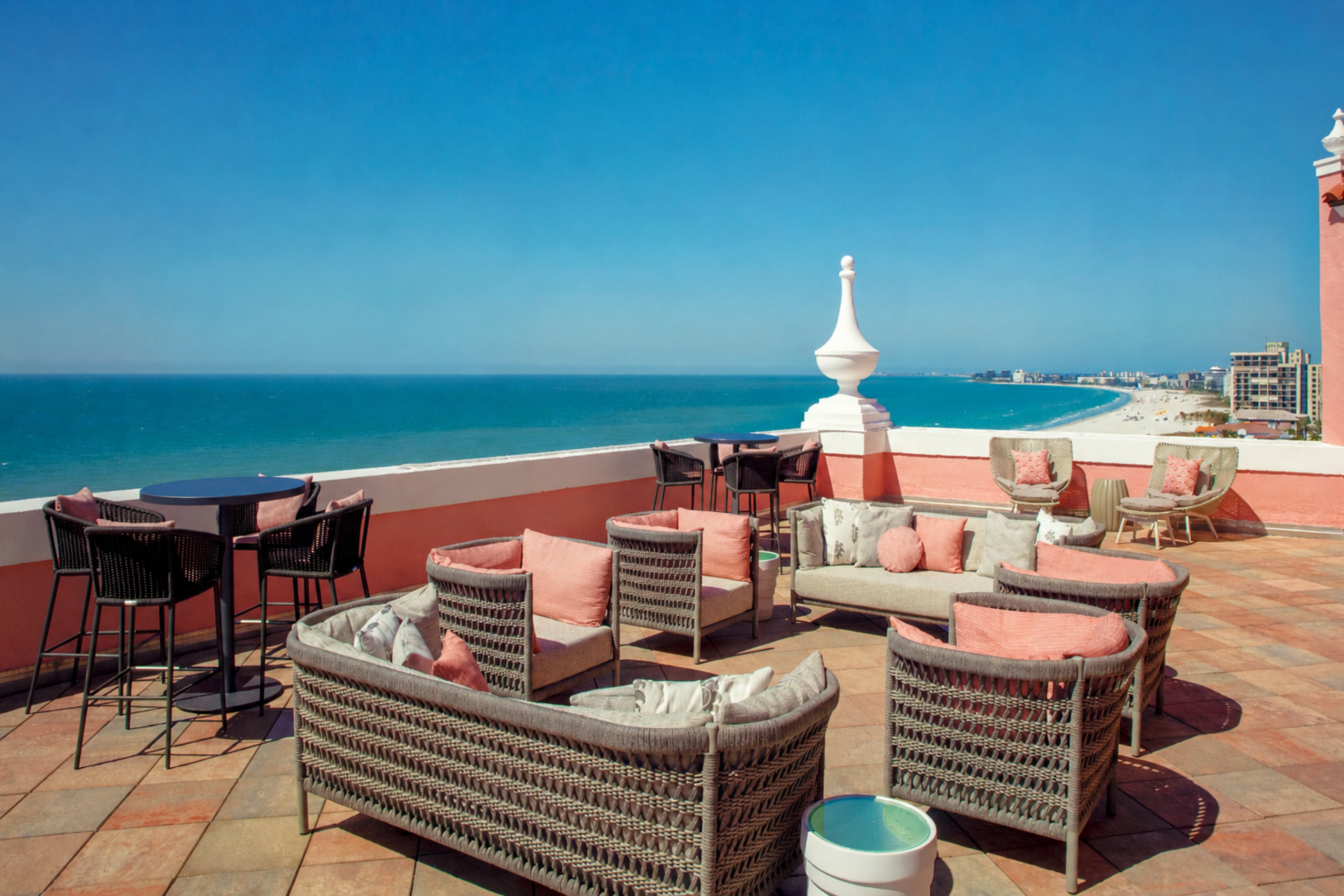 Seaside rooftop lounge with wicker chairs and pink cushions overlooking a blue ocean.