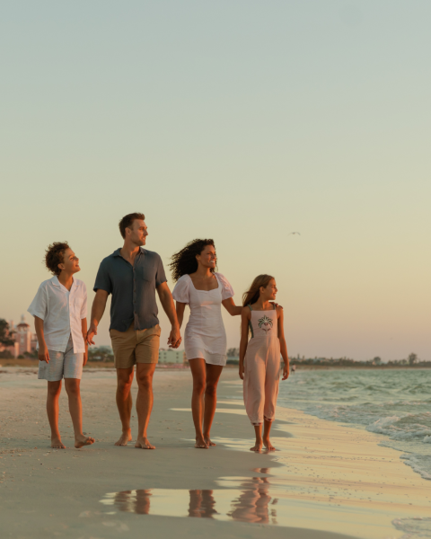 Family of four walking along the beach hand-in-hand at sunset, waves at their feet.