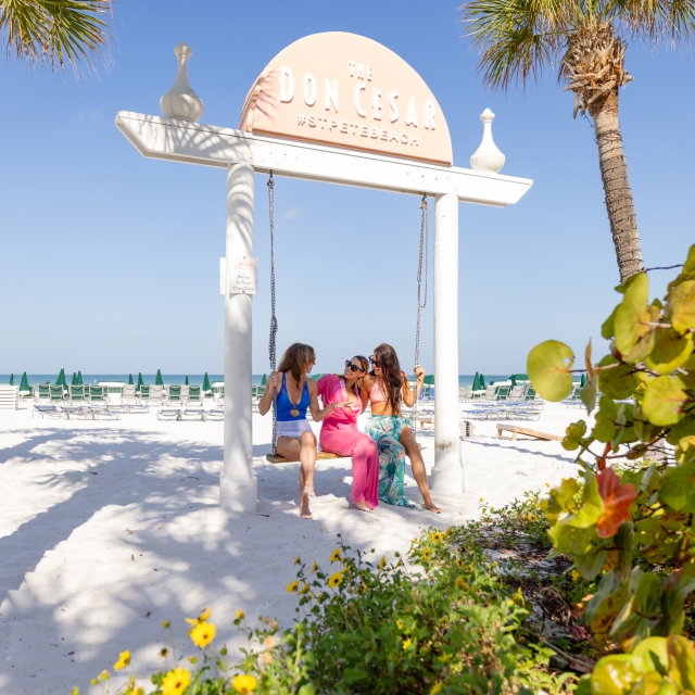 Women laughing and relaxing on a swing at the beach, with the ocean in the background and a clear blue sky above.