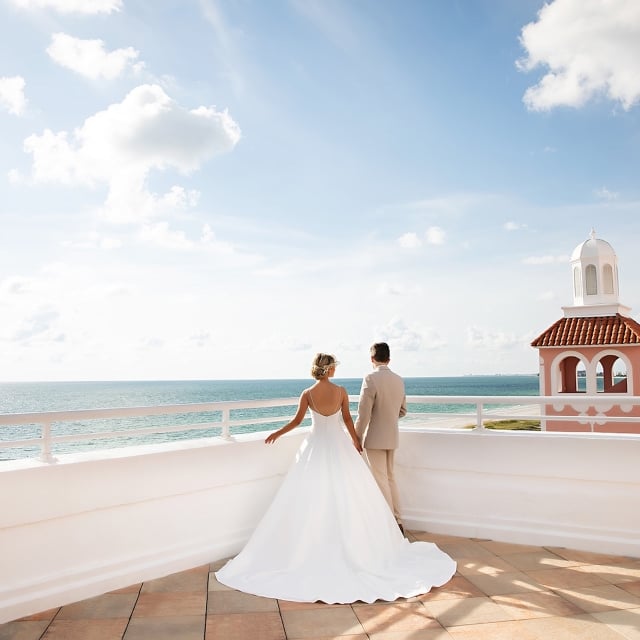 A bride and groom stand on a balcony, gazing at the ocean, surrounded by a romantic atmosphere.