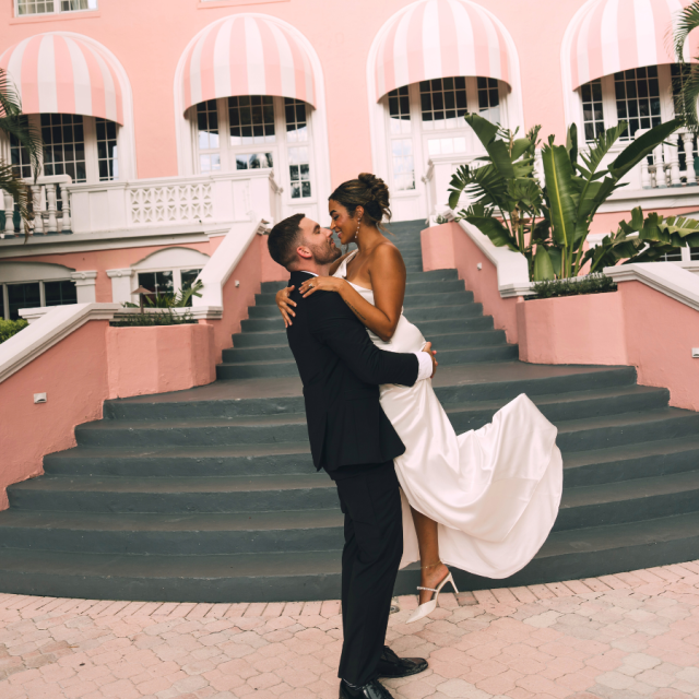 A bride and groom share a loving embrace in front of a charming pink hotel, capturing a moment of joy and celebration.