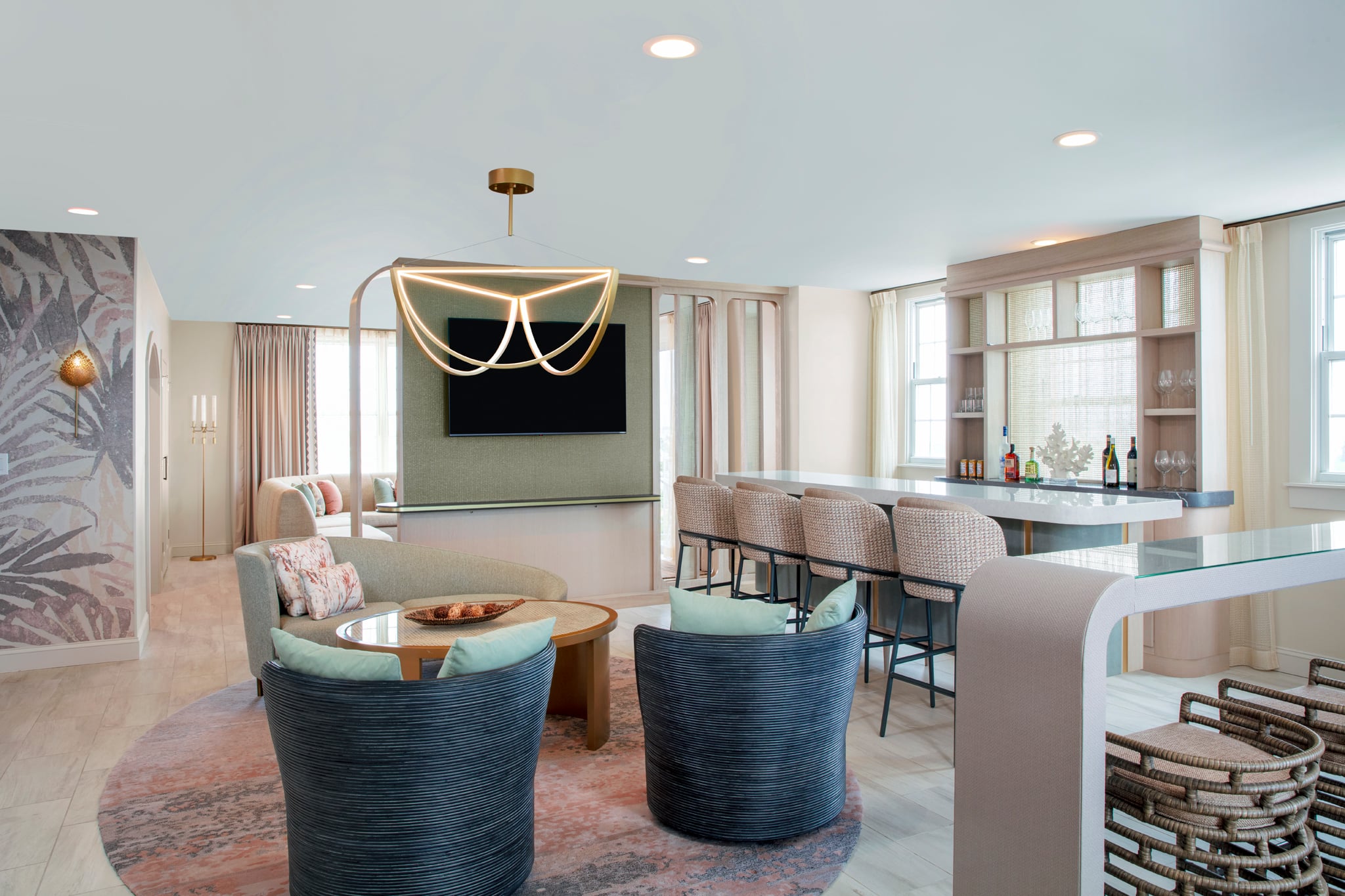 Open-concept living area with a modern chandelier, curved black woven chairs around a glass coffee table, and a bar with bottles along a windowed wall.