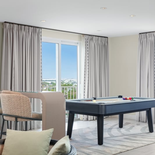 Bright living room with beige wicker chairs around a black pool table, light gray curtains, and large windows showing a coastal city view.