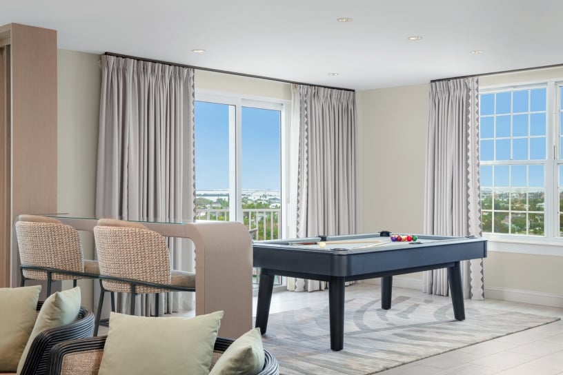 Bright living room with beige wicker chairs around a black pool table, light gray curtains, and large windows showing a coastal city view.