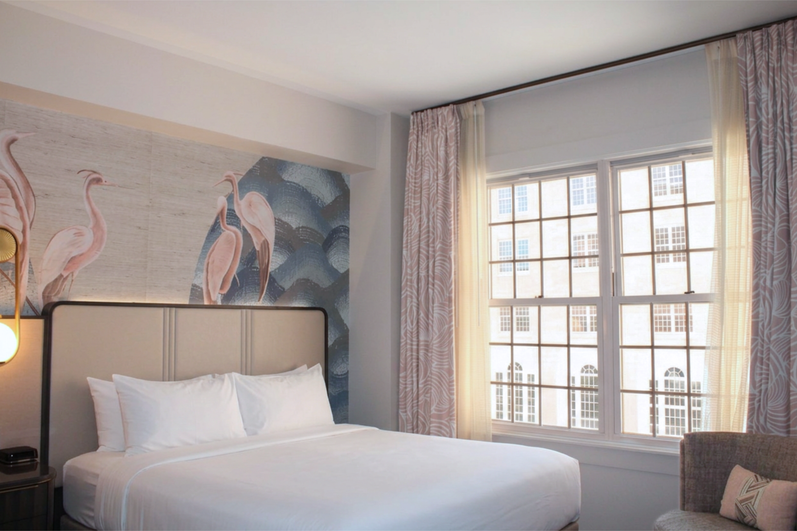Hotel bedroom with a white bed and padded headboard, a pink flamingo mural on the wall, and a large window with sheer and patterned curtains letting in daylight.
