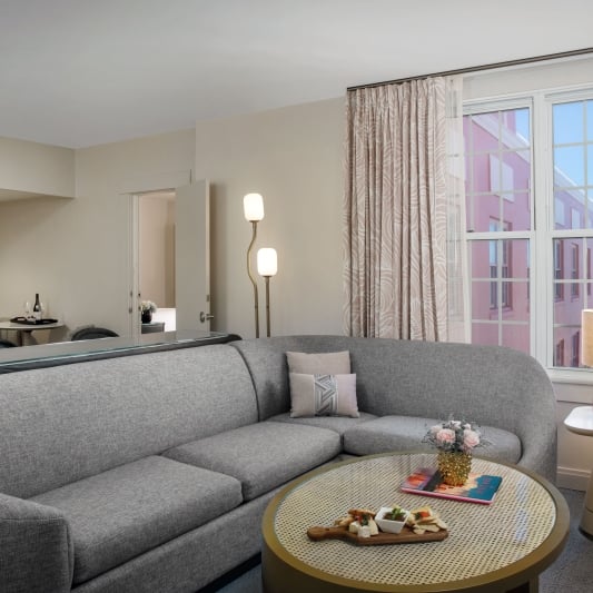 Living room with a gray sectional sofa, round perforated coffee table with snacks, and large windows showing a pink building outside.
