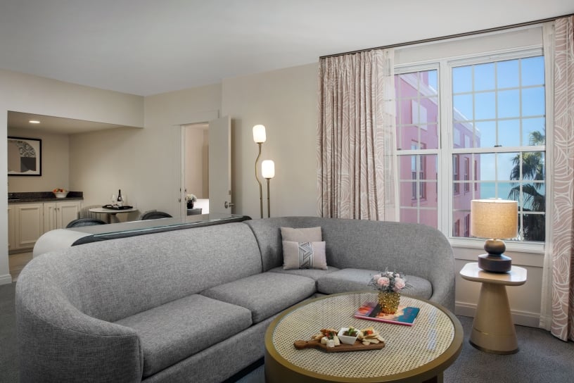 Living room with a gray sectional sofa, round perforated coffee table with snacks, and large windows showing a pink building outside.