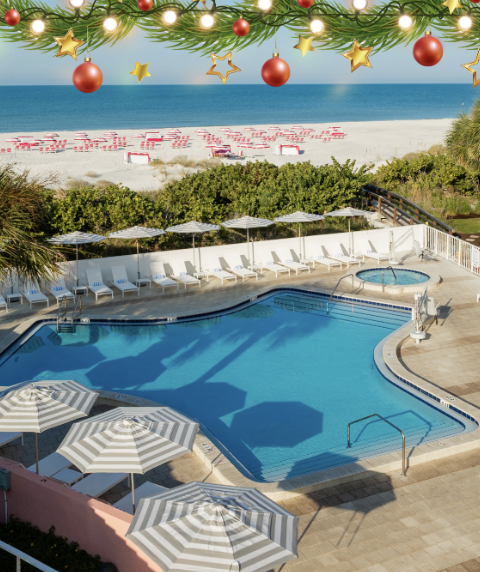 Resort pool area with white loungers and striped umbrellas, palm trees, pink building, and a sunny beach beyond, with festive garlands and ornaments overhead.