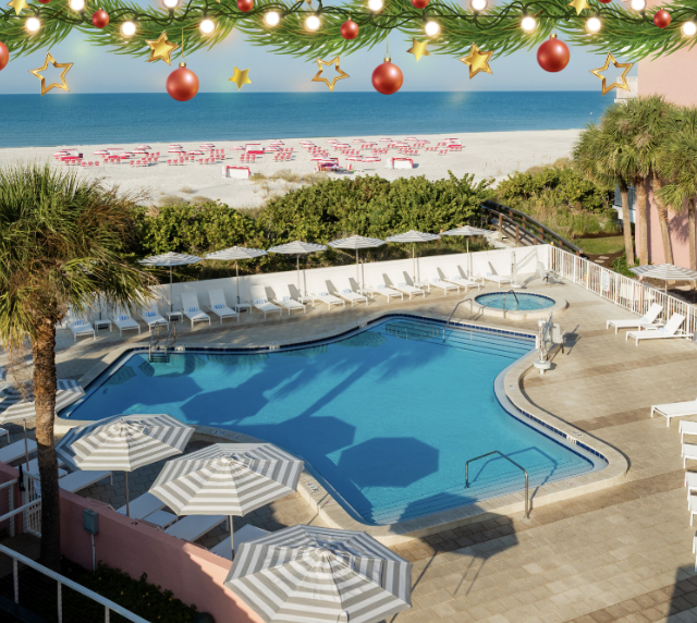 Resort pool area with white loungers and striped umbrellas, palm trees, pink building, and a sunny beach beyond, with festive garlands and ornaments overhead.