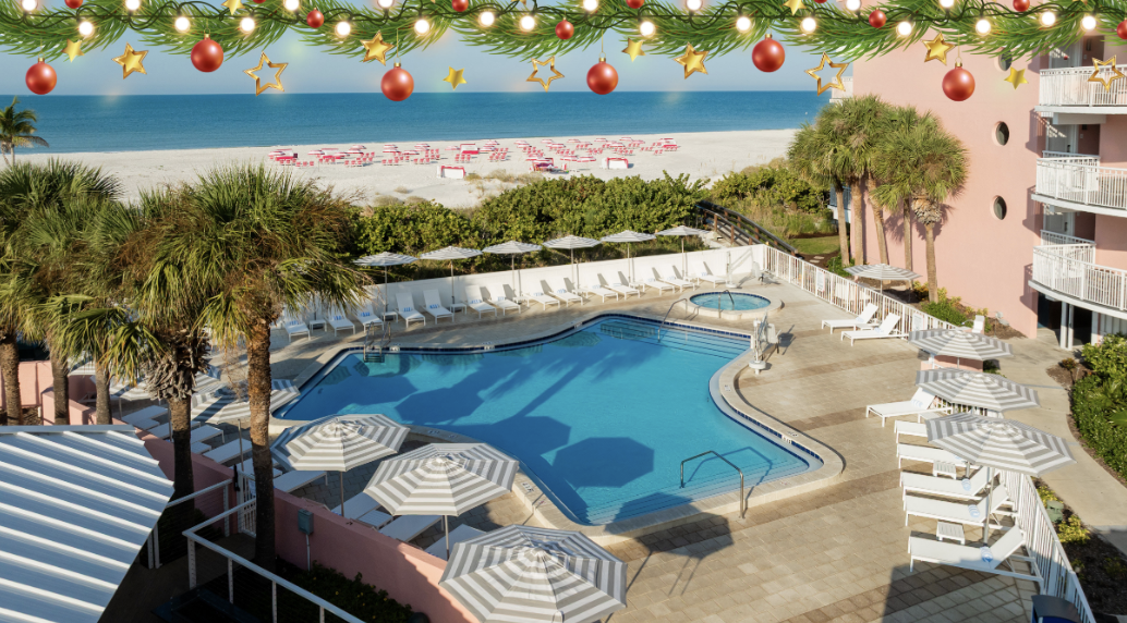 Resort pool area with white loungers and striped umbrellas, palm trees, pink building, and a sunny beach beyond, with festive garlands and ornaments overhead.
