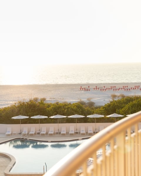 Hotel balcony railing in the foreground overlooking a beach and ocean at sunset, with a pool and white lounge chairs below and palm trees nearby.