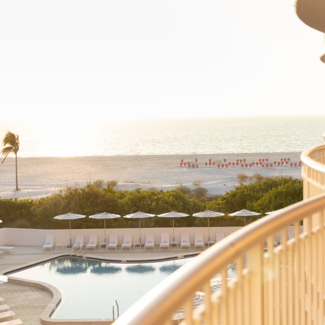 Hotel balcony railing in the foreground overlooking a beach and ocean at sunset, with a pool and white lounge chairs below and palm trees nearby.