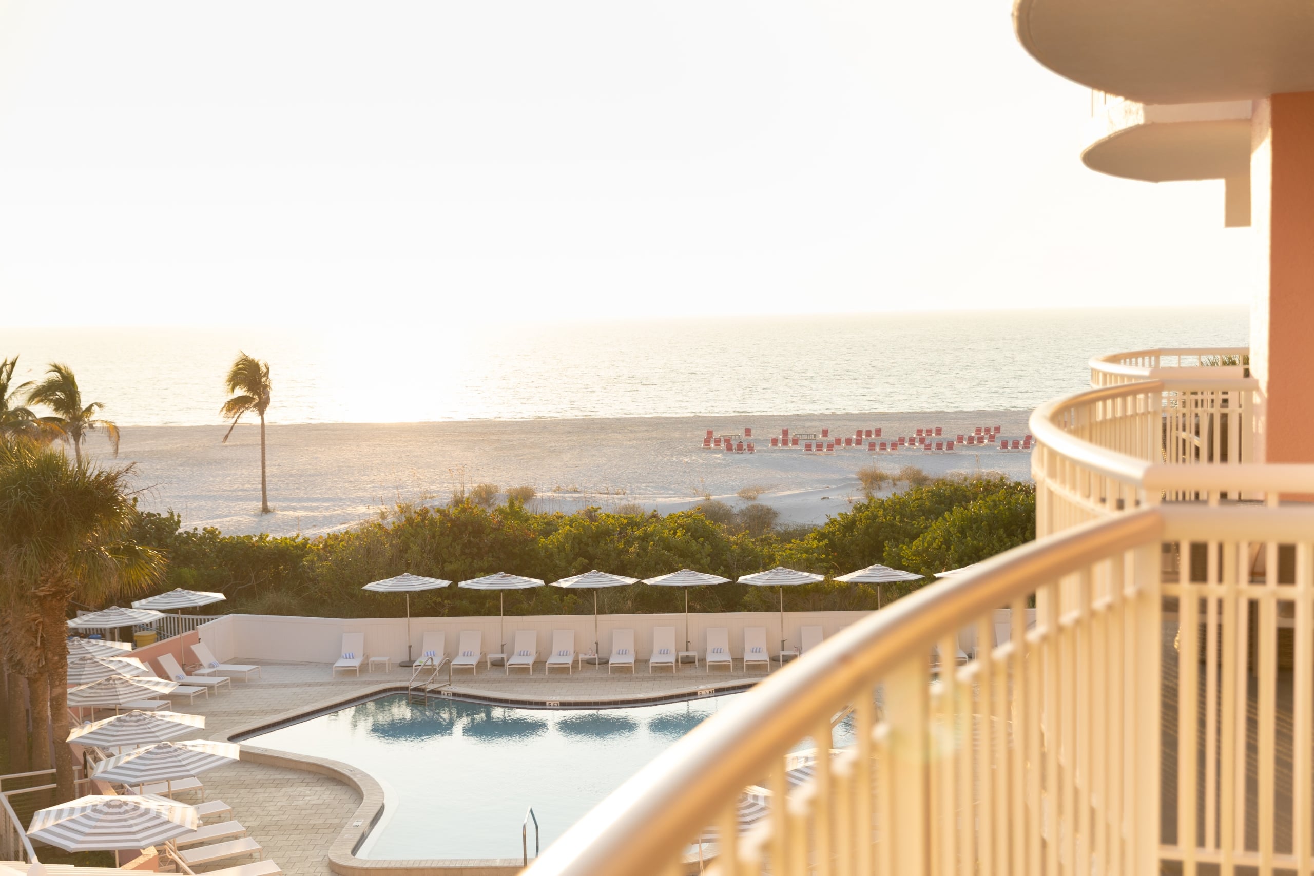 Hotel balcony railing in the foreground overlooking a beach and ocean at sunset, with a pool and white lounge chairs below and palm trees nearby.