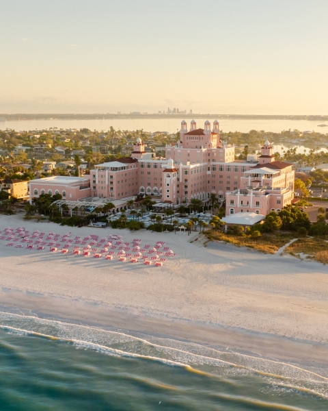 Aerial view of a pink hotel on a sandy beach with pink umbrellas and turquoise water in the foreground.