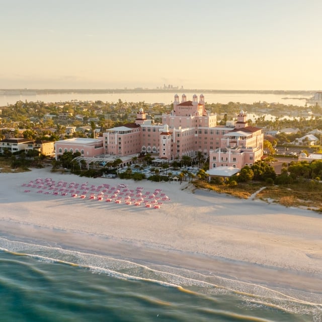 Aerial view of a pink hotel on a sandy beach with pink umbrellas and turquoise water in the foreground.