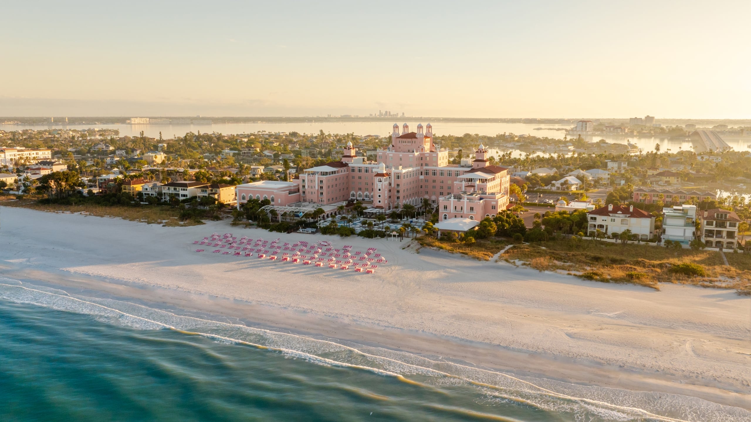 Aerial view of a pink hotel on a sandy beach with pink umbrellas and turquoise water in the foreground.
