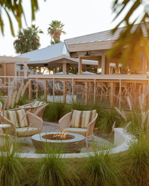 Sunlit outdoor lounge with wicker chairs around a fire pit and tall ornamental grasses.