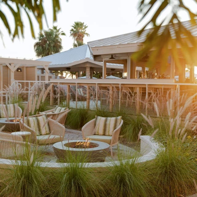 Sunlit outdoor lounge with wicker chairs around a fire pit and tall ornamental grasses.