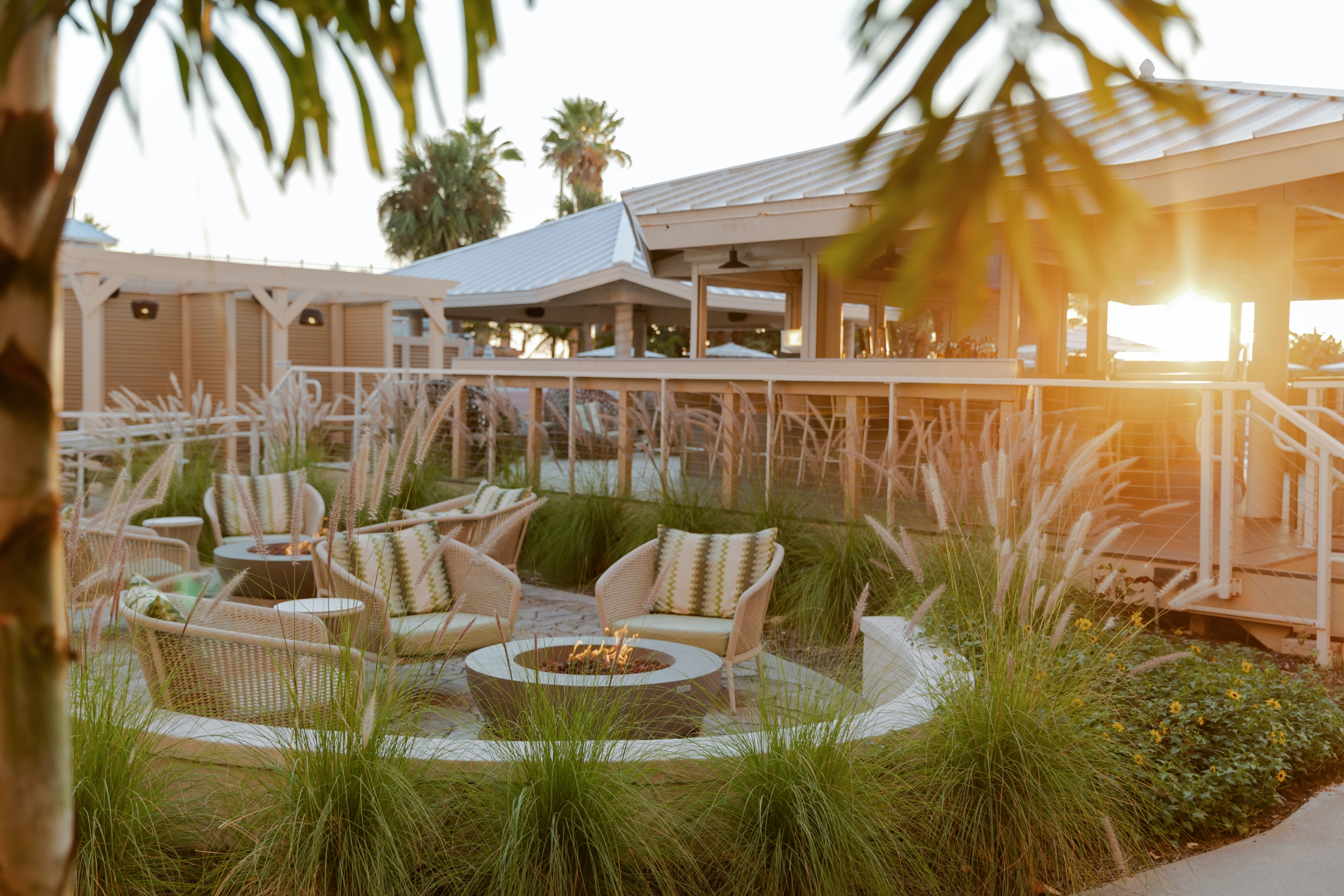 Sunlit outdoor lounge with wicker chairs around a fire pit and tall ornamental grasses.
