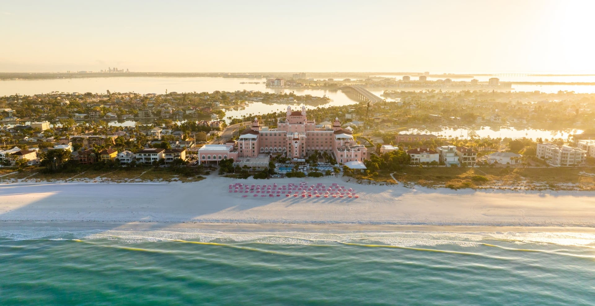Aerial view of a pink resort hotel on a sandy beach with rows of pink umbrellas and a calm ocean beyond, at sunset.