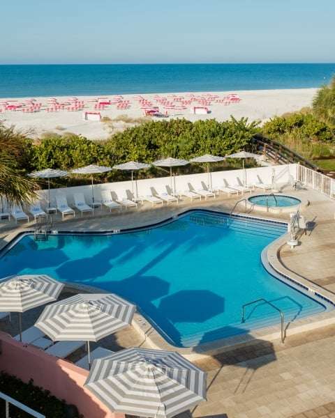 Resort pool area with white lounge chairs, striped umbrellas, and palm trees; beach and blue ocean beyond.
