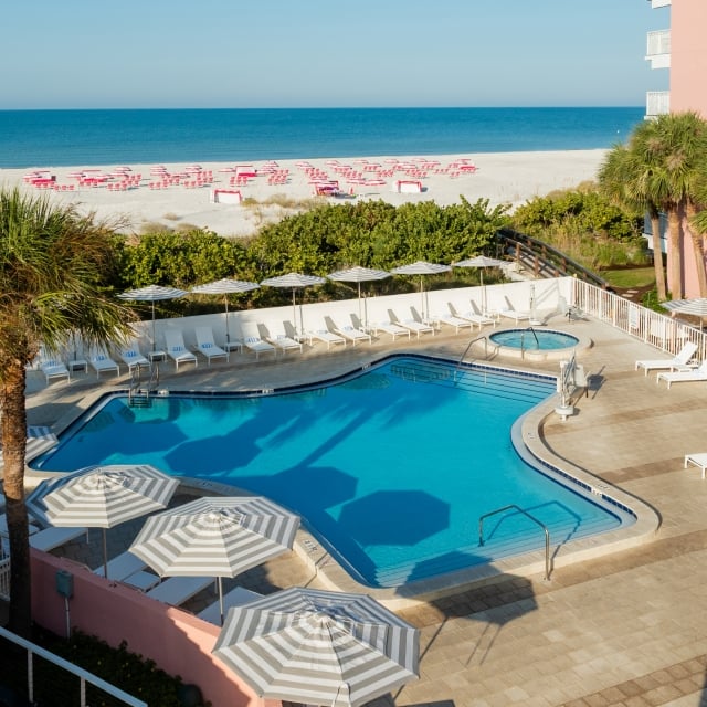 Resort pool area with white lounge chairs, striped umbrellas, and palm trees; beach and blue ocean beyond.