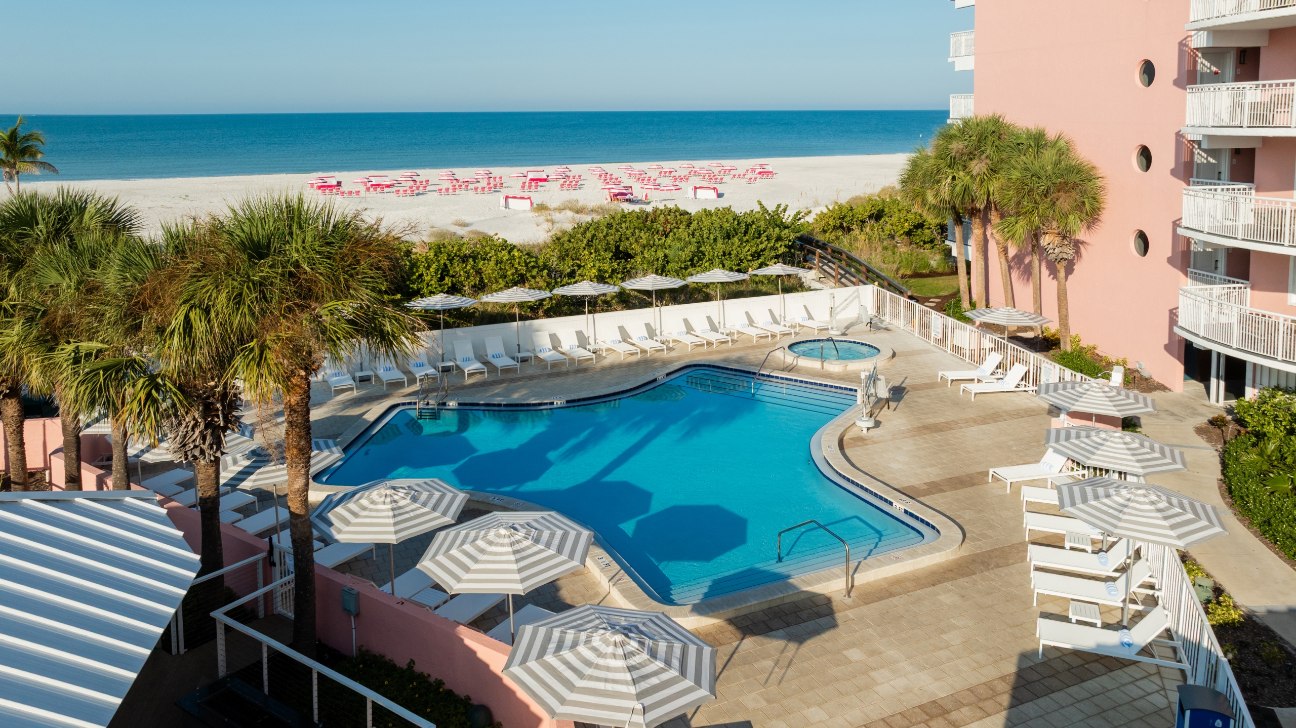 Resort pool area with white lounge chairs, striped umbrellas, and palm trees; beach and blue ocean beyond.