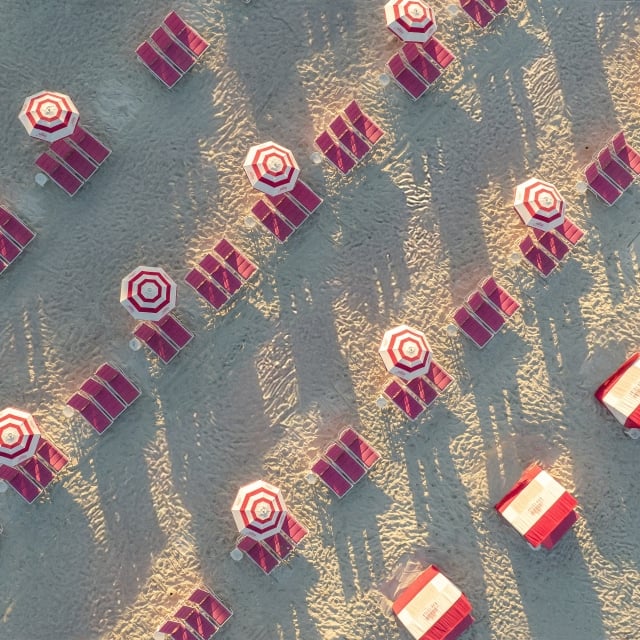 Aerial view of red and white striped umbrellas with matching lounge chairs on a sandy beach, arranged in rows with long shadows.