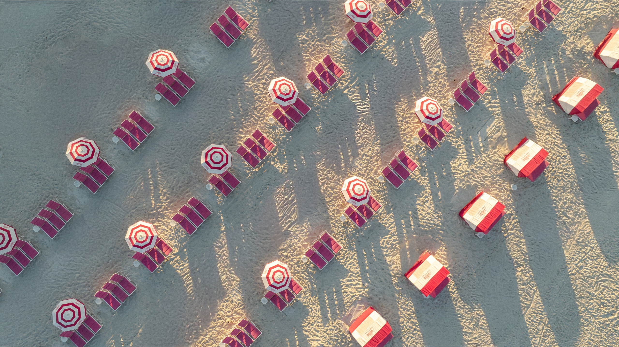 Aerial view of red and white striped umbrellas with matching lounge chairs on a sandy beach, arranged in rows with long shadows.